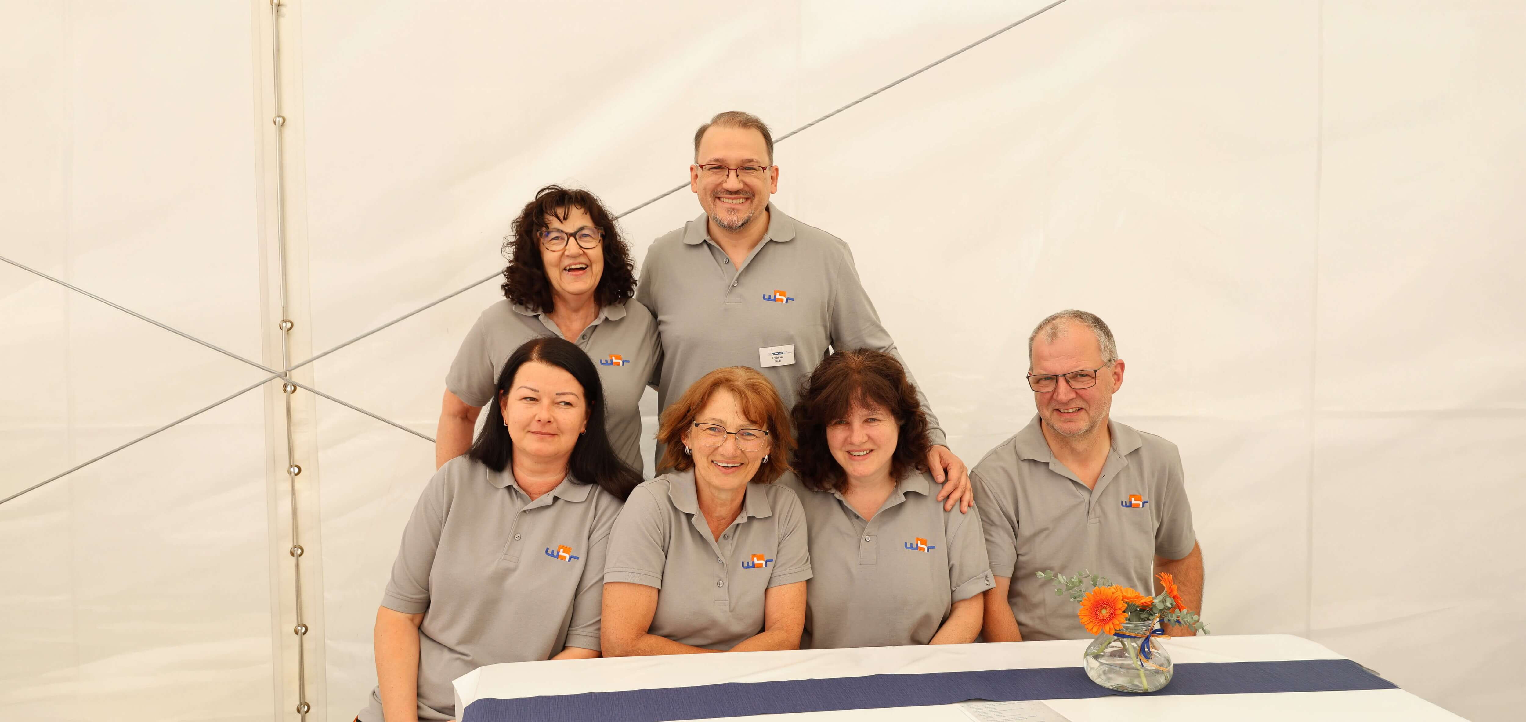 A friendly group of six employees wearing grey shirts smiles together at a table adorned with a small flower vase in a tent setting.